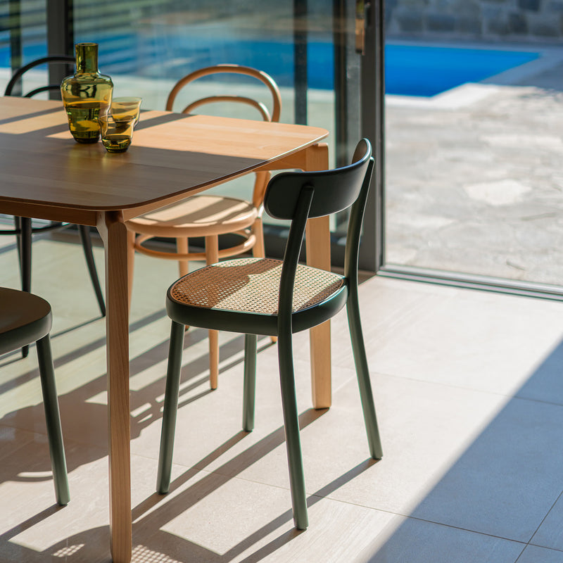 Sunlit dining area with a solid wood table and TONs Chair La Zitta - Cane, featuring ergonomic woven rattan seats. Two green glass vases sit on the table, while large glass doors provide a view of the outdoor pool and stone patio.