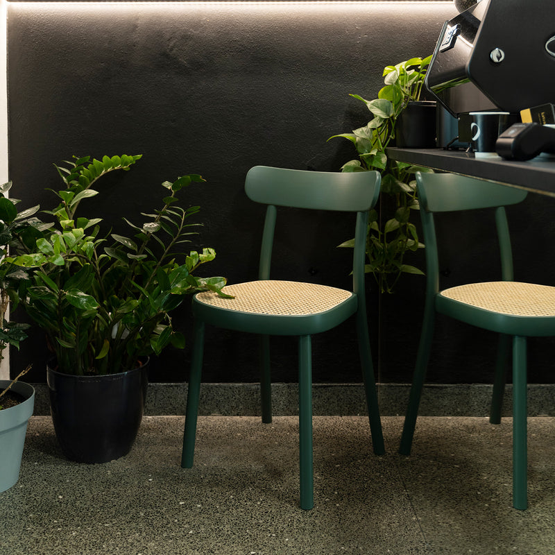Two green Chair La Zitta - Cane by TON, ergonomically designed, create a cozy corner next to a coffee machine on a counter. Potted plants surround the chairs, adding greenery against the dark wall, subtly lit from above.