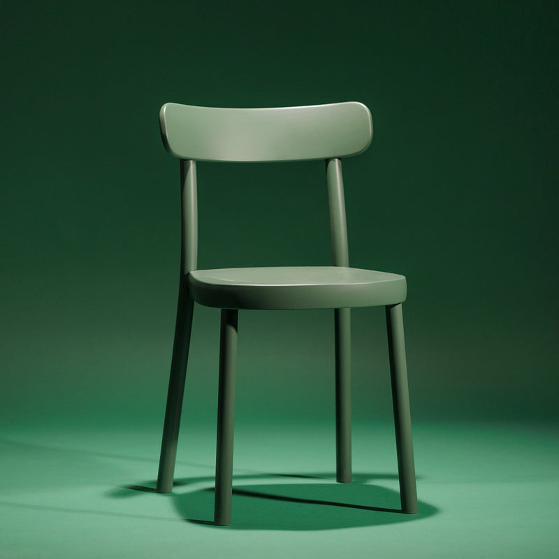 The Chair La Zitta from TON is crafted in classic bentwood style, featuring a curved backrest and four straight legs. Its showcased against a matching green background.