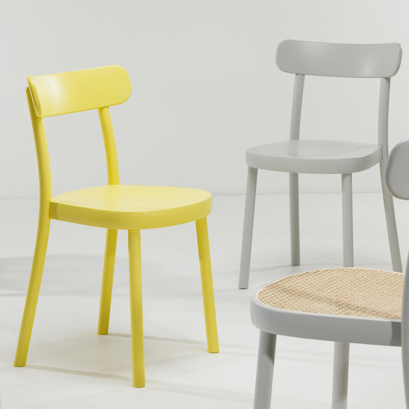 A minimalist scene features Alex Guflers creations for TON: the bright yellow and light gray Chair La Zitta with solid wood frames, plus a gray model with an ergonomic rattan seat, all elegantly showcased on a pristine white floor against a plain backdrop.