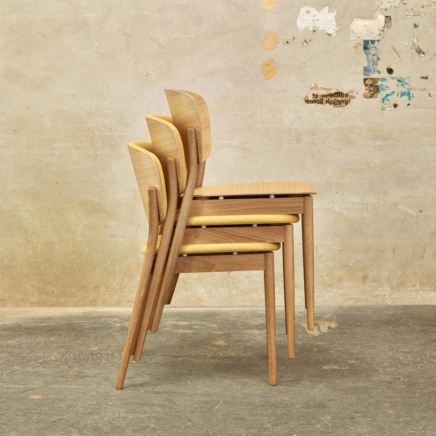 Three Chair Valencia dining chairs by TON, featuring solid wood construction with light brown seats and backs and slender legs, are stacked against a textured, paint-scratched wall on an aged concrete floor for a minimalist look.