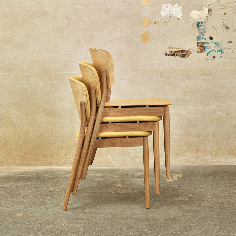 Three Chair Valencia dining chairs by TON, featuring solid wood construction with light brown seats and backs and slender legs, are stacked against a textured, paint-scratched wall on an aged concrete floor for a minimalist look.