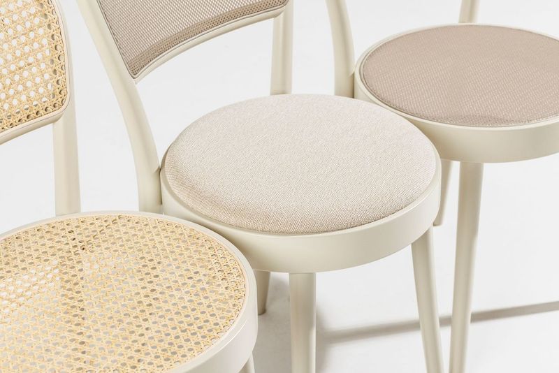A close-up showcases four TON Chair 314 chairs by TON with round seats and cream-colored frames. Two have woven cane surfaces, and two feature light beige upholstery. The chairs, featuring bentwood craftsmanship, are arranged in a staggered formation for visual interest.