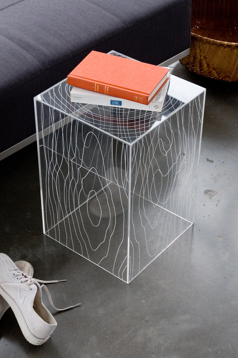 Transparent cube side table with white wood grain pattern holding two books beside dark gray sofa on concrete floor