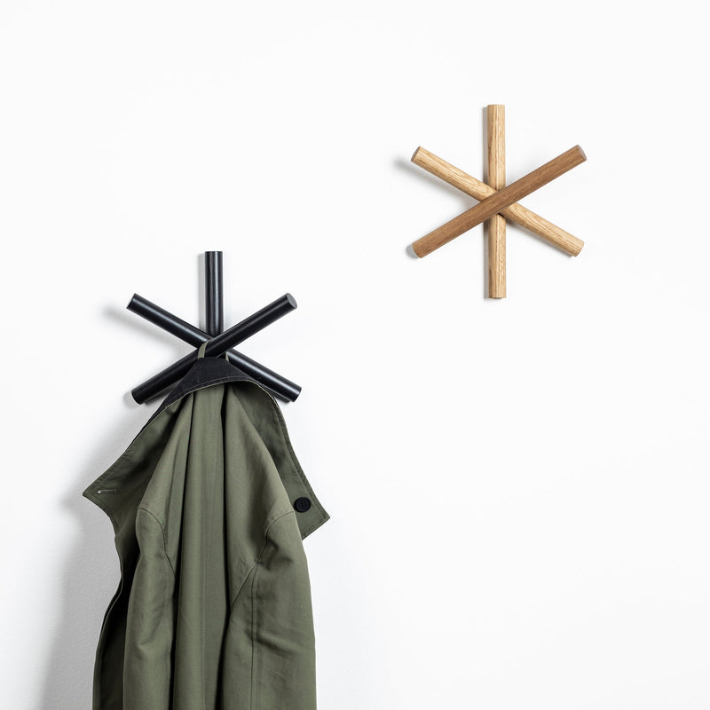 A green jacket hangs on a black, asterisk-shaped wall hook. Nearby, the Logs - Inventory hook by TON, crafted from quality wood offcuts, stands empty. Both hooks are mounted on a white wall.