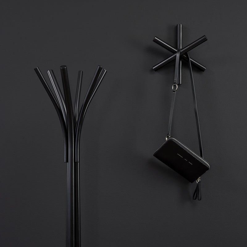 Next to a wall-mounted asterisk-shaped rack, a minimalist black coat rack crafted from high-quality wood offcuts stands. A small black bag hangs on the wall rack against a dark gray background, highlighting sustainable design principles. Featured product: Logs - Inventory by TON.