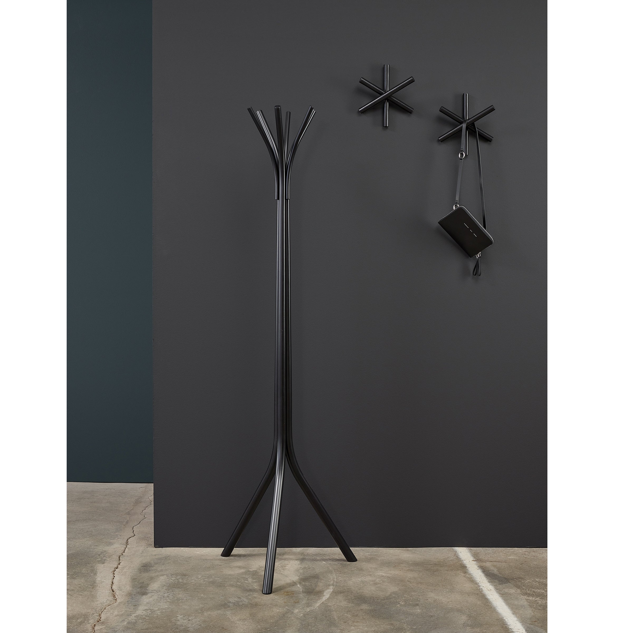 A minimalist room features a stylish modern black coat rack and two unique wall hooks—crafted from sustainable wood offcuts by TON—against a dark wall. One hook elegantly holds a small black bag, while the concrete floor complements the contemporary aesthetic of the design.