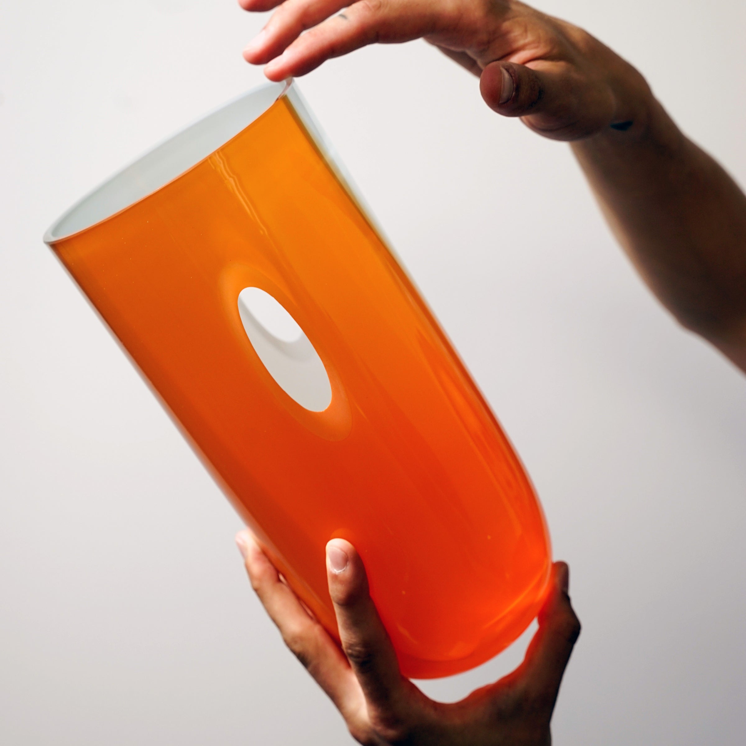 Hands delicately hold Ursula Futuras ECLIPSE Vase, characterized by its tall, cylindrical shape and central oval cutout. Its glossy finish suggests mouth-blown glass or ceramic craftsmanship against a plain backdrop.