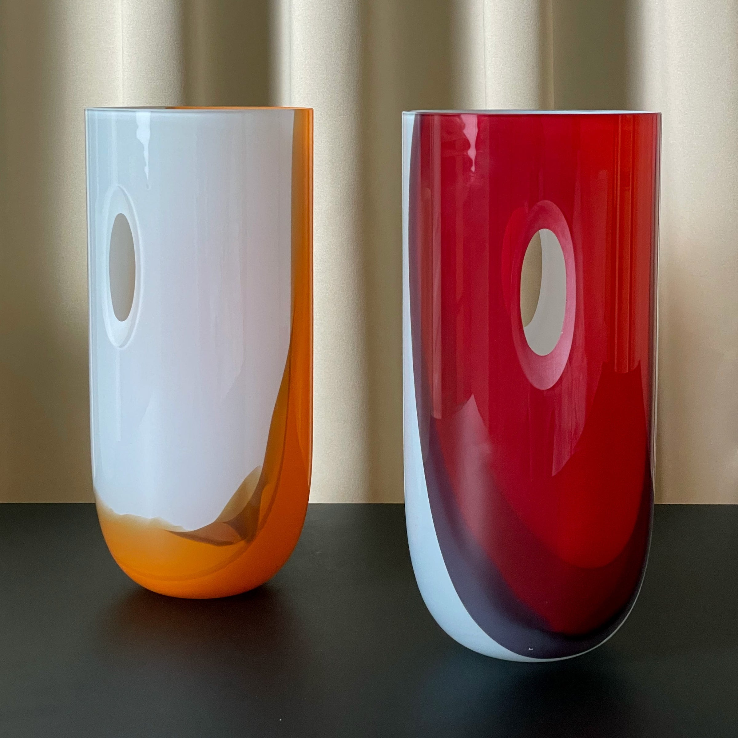 Two elongated ECLIPSE vases from Ursula Futura rest on a black surface against a gold backdrop. The left one, mouth-blown glass with white and orange accents, the right with red accents. Both feature a unique design and an oval cutout near the top.