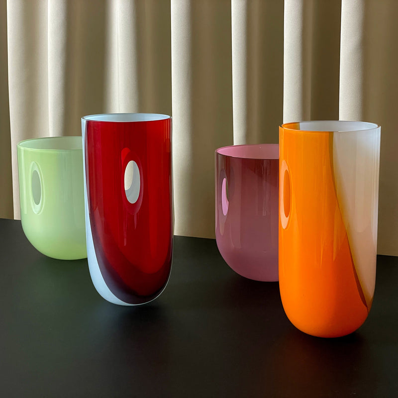Four colorful ECLIPSE Vases by Ursula Futura, in green, red, pink, and orange with oval openings, are set on a black surface against a beige curtain. Crafted from mouth-blown glass with a glossy finish and curved design, these modern vases offer a unique look.