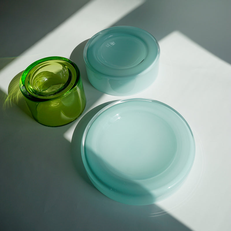 In sunlight, two WET Bowl Big dishes by Ursula Futura, smooth and translucent in their blue-green hue, rest on a white surface alongside a taller green cylindrical container. Soft shadows and reflections enhance their artistic allure.