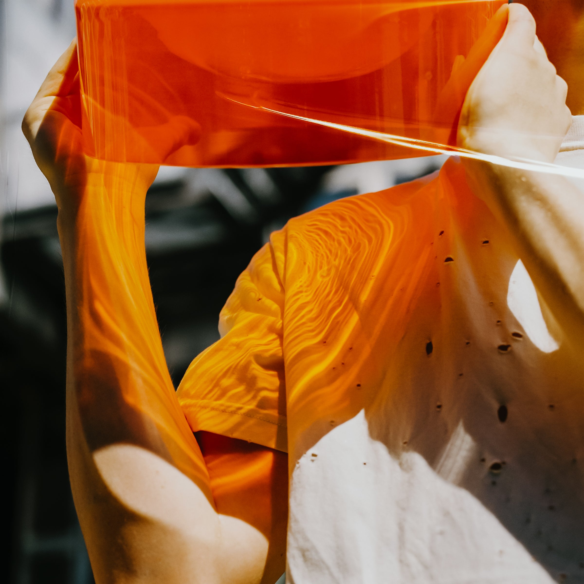 Someone holds up an orange translucent sheet, reminiscent of the WET Bowl Big by Ursula Futura, against a bright background. Light filters through, casting warm, wavy patterns on their white shirt and skin, creating an abstract and artistic scene with contrasting textures and tones.