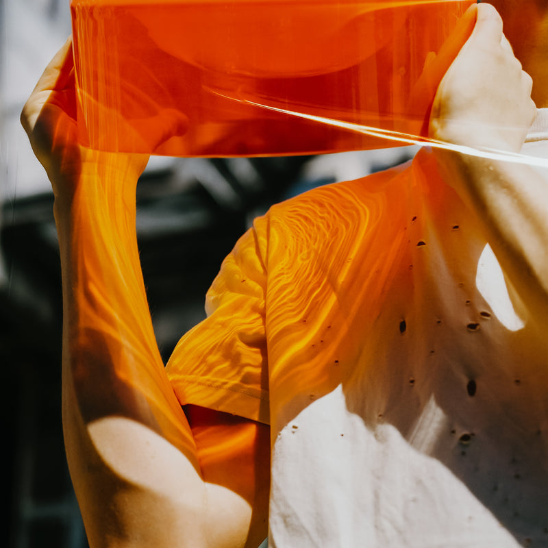 Someone holds up an orange translucent sheet, reminiscent of the WET Bowl Big by Ursula Futura, against a bright background. Light filters through, casting warm, wavy patterns on their white shirt and skin, creating an abstract and artistic scene with contrasting textures and tones.