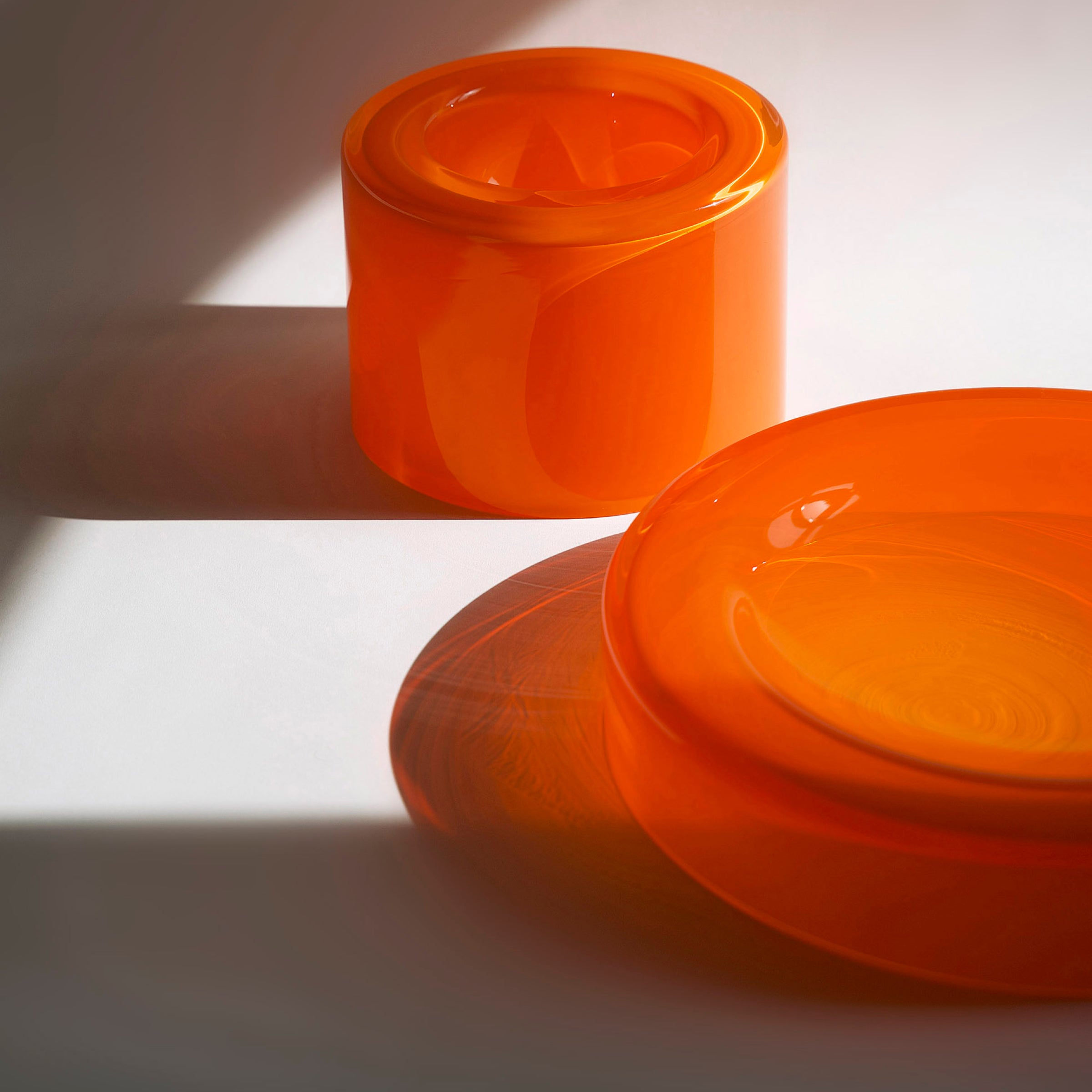 Two vibrant orange glass objects attract attention on a light surface: one is a cylindrical piece, and the other is the WET Bowl Big by Ursula Futura, which radiates charm. Both cast soft shadows as the lighting highlights their glossy texture and rich color.