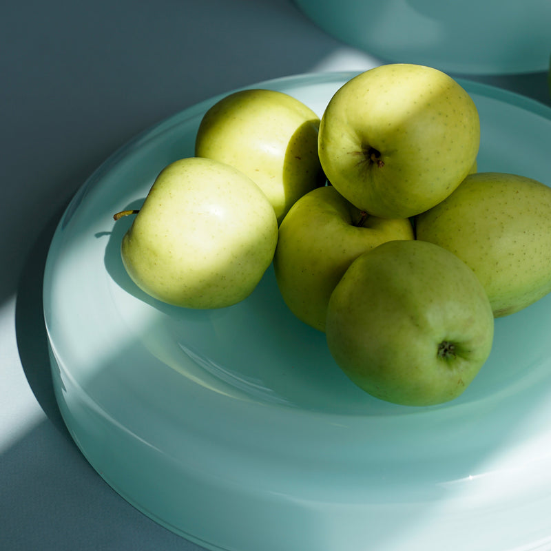 Six green apples are elegantly arranged on a mouth-blown WET Bowl Big by Ursula Futura. Sunlight casts soft shadows, enhancing their shine against a calm, minimalist blue-grey backdrop.