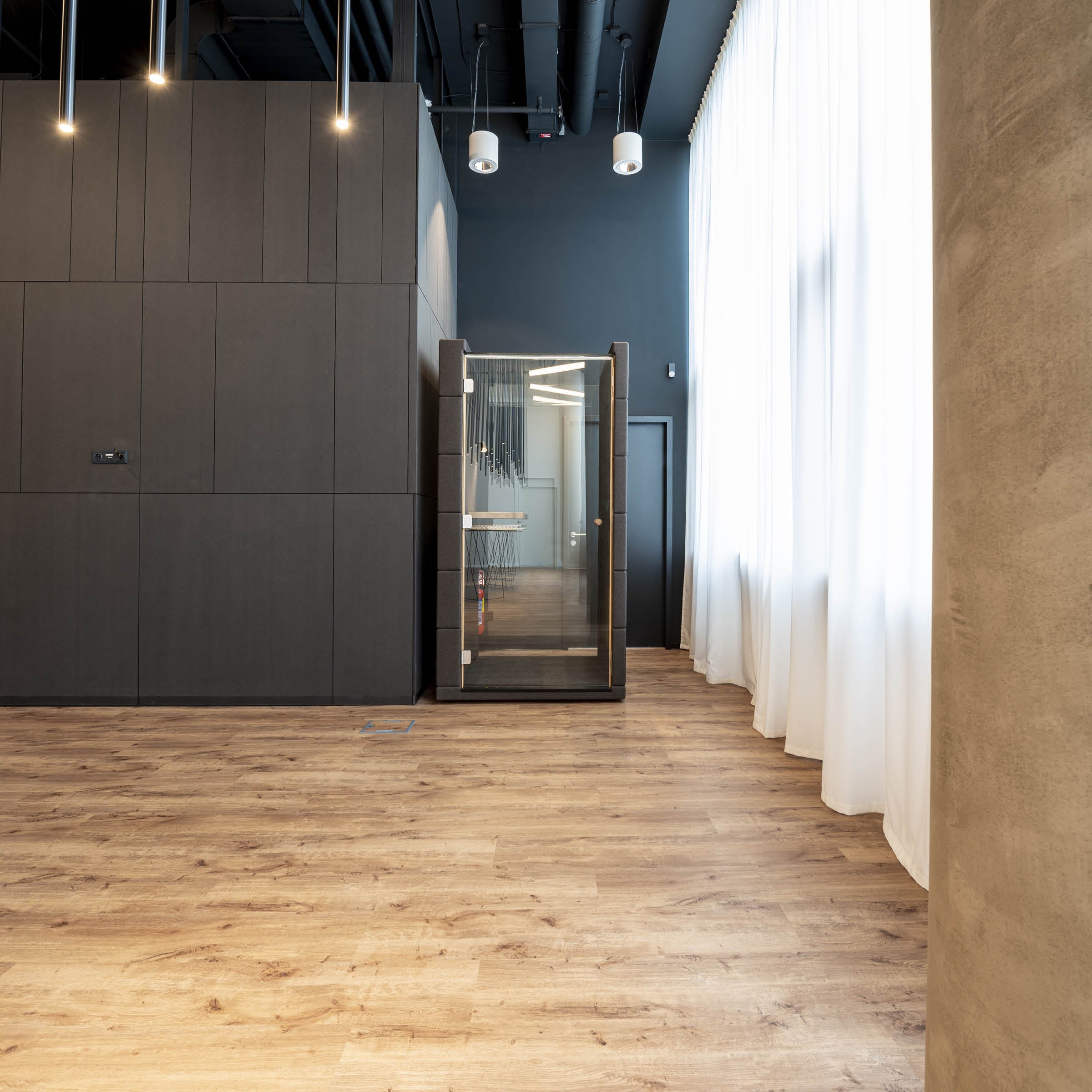 The OFFICE POD UNIQ by SilentLab boasts modern minimalist décor with wooden flooring, tall white curtains, and dark walls. A sleek glass door reveals a compact exercise space with gym gear. Ceiling lights brighten the room, enhanced by soundproofing for a serene experience.