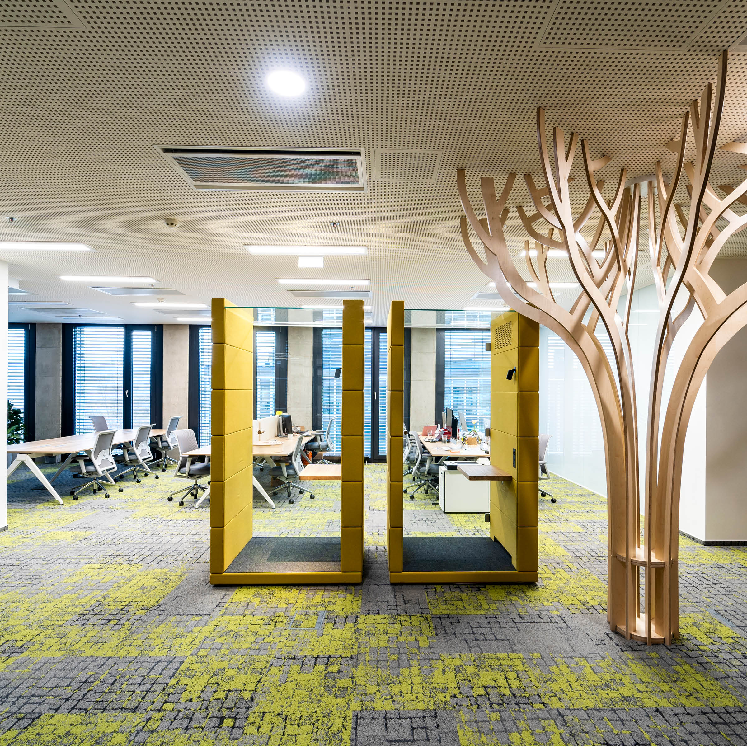 The OFFICE POD UNIQ by SilentLab offers a modern space with open workstations, vibrant green and gray carpet, yellow dividers, and a unique tree-like wooden structure. Large windows provide natural light, while acoustic upholstery ensures soundproofing for enhanced focus.