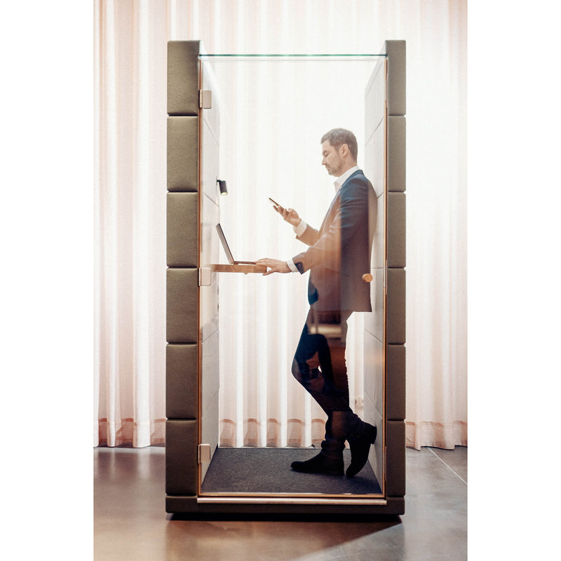 Dressed in business attire, a person stands inside SilentLabs OFFICE POD UNIQ, a modern glass-walled booth with acoustic upholstery for soundproofing. They work on a laptop and check their phone in a softly lit room with sheer curtains in the background.