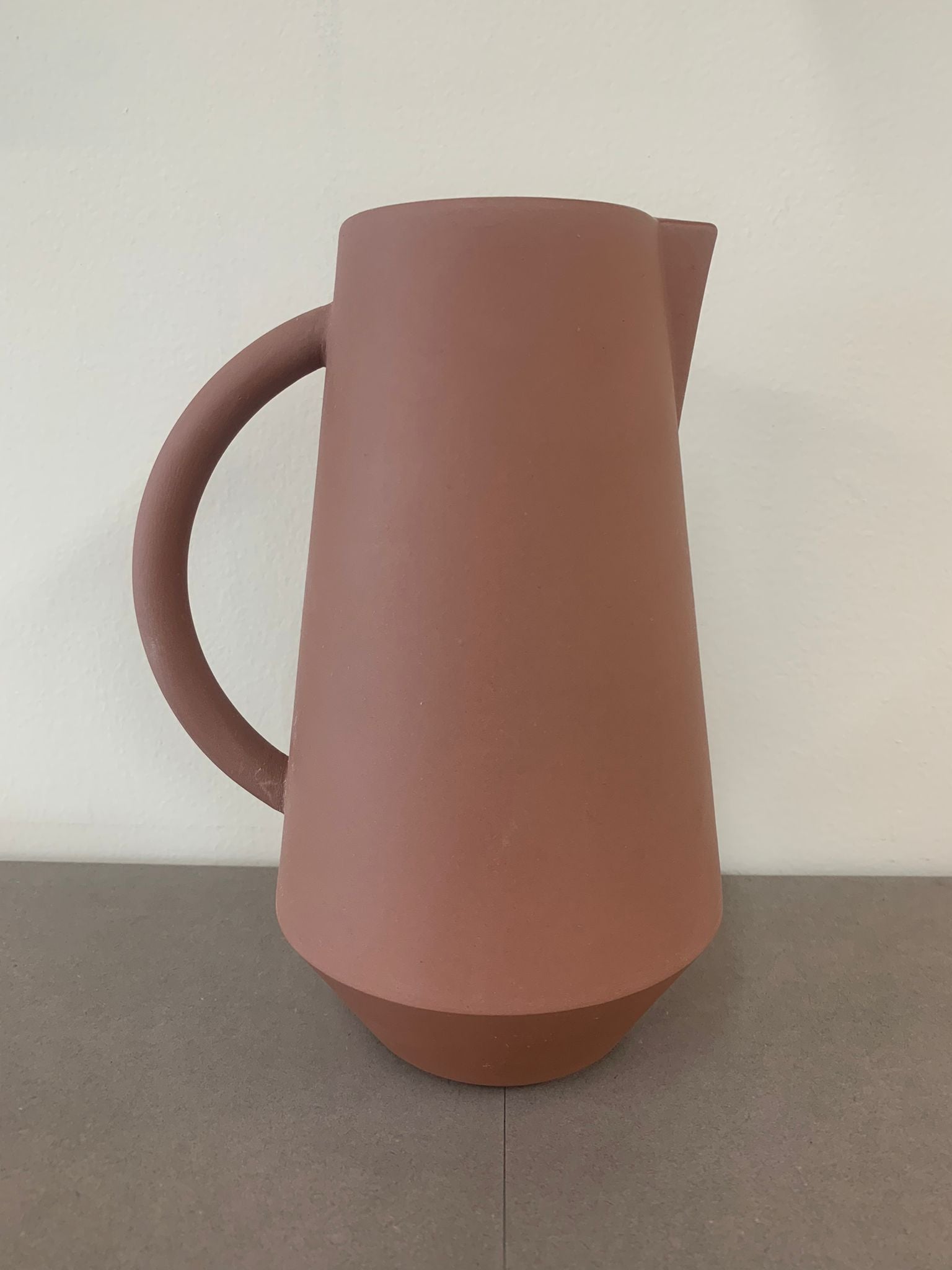 The Unison Carafe by Schneid, a part of their stackable ceramic tableware handmade in Germany, is a matte, terracotta-colored pitcher with a wide base, narrow neck, and curved handle. It elegantly sits on a gray surface against a light background.