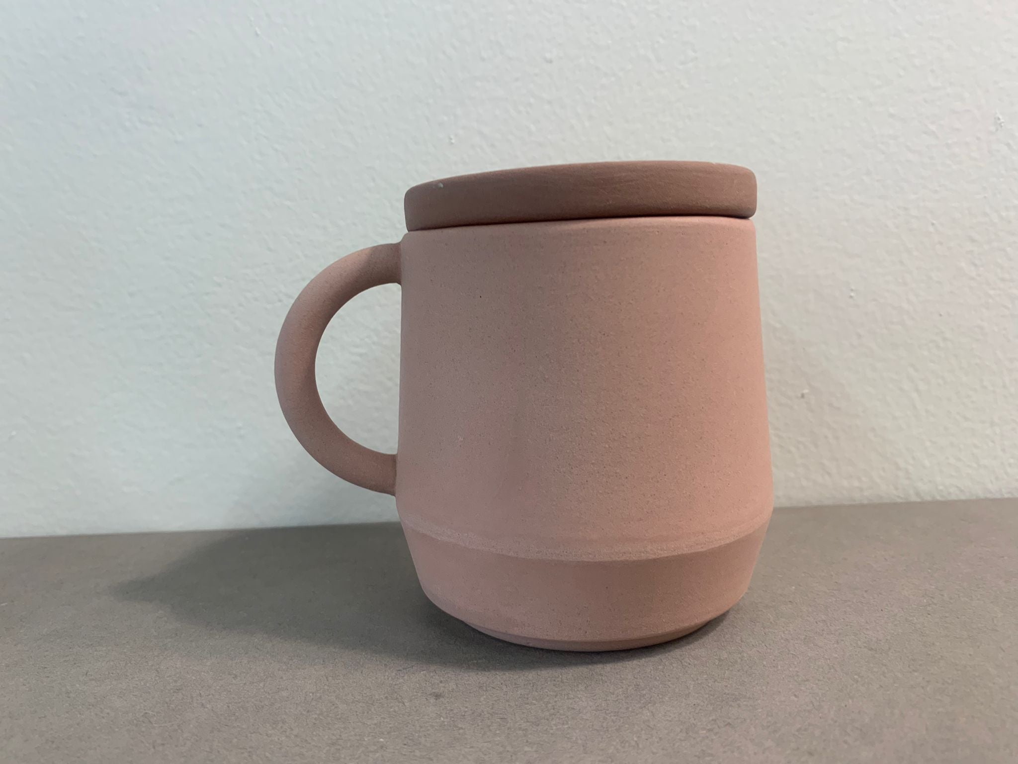 The Unison Cup - Sale by Schneid is a terracotta matte ceramic mug with a matching lid, set on a gray surface. Featuring a simple handle and smooth unglazed finish against an off-white background, its stackable design adds practicality and elegance.