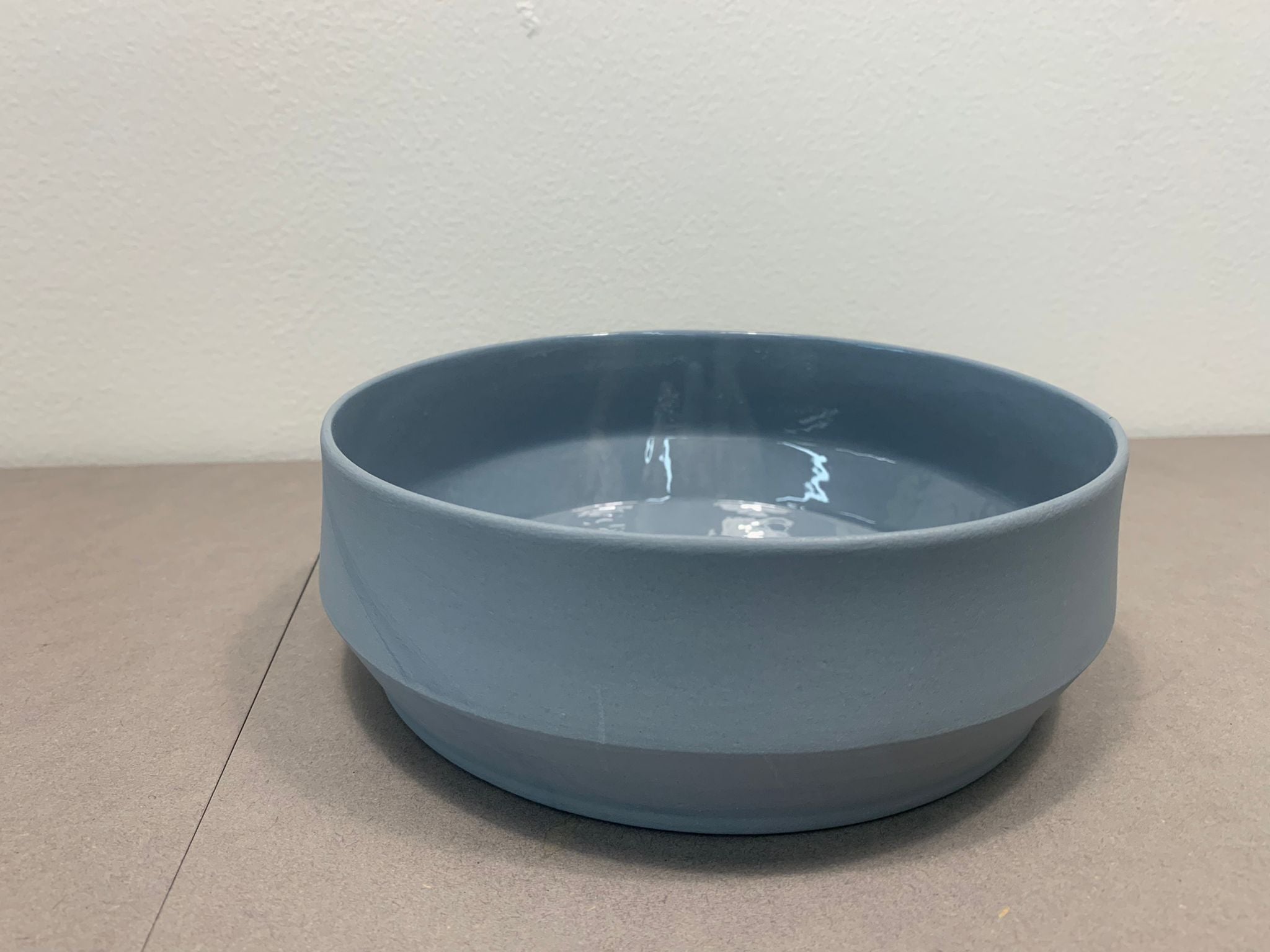 Placed on a neutral gray surface against a plain white wall, the Unison Soup Bowl by Schneid features a simple, matte blue ceramic design and is handmade in Germany.