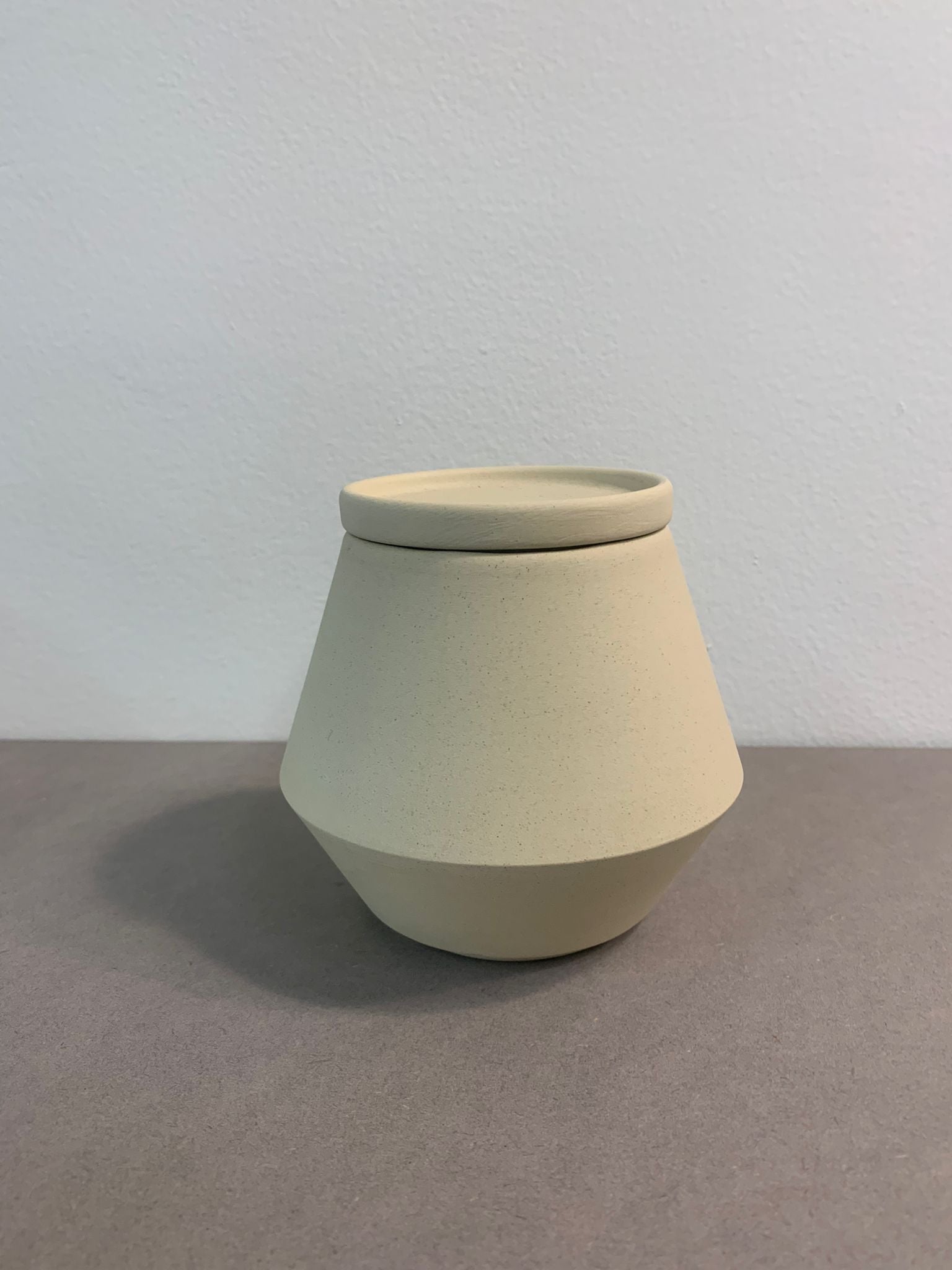 The beige Unison Sugar Bowl by Schneid, a geometric ceramic jar with a conical shape and lid, from their sophisticated tableware collection, rests on a gray surface against a light gray wall.