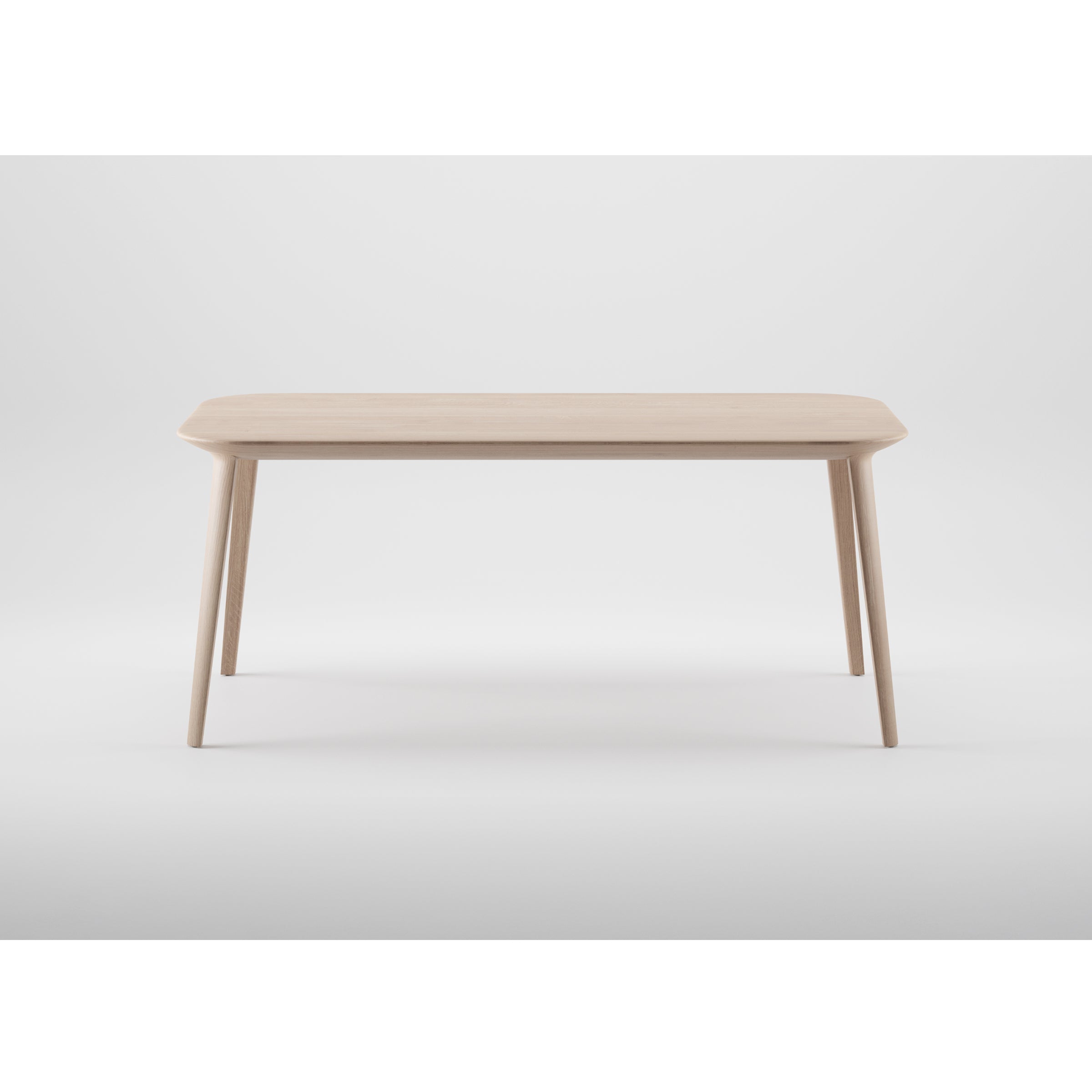 The KALOTA Table by Artisan is a minimalist wooden table crafted from solid wood with a light finish. It features a rectangular top with rounded edges and four slender, tapering legs, all finished with natural oils, creating an elegant design set against a plain white background.