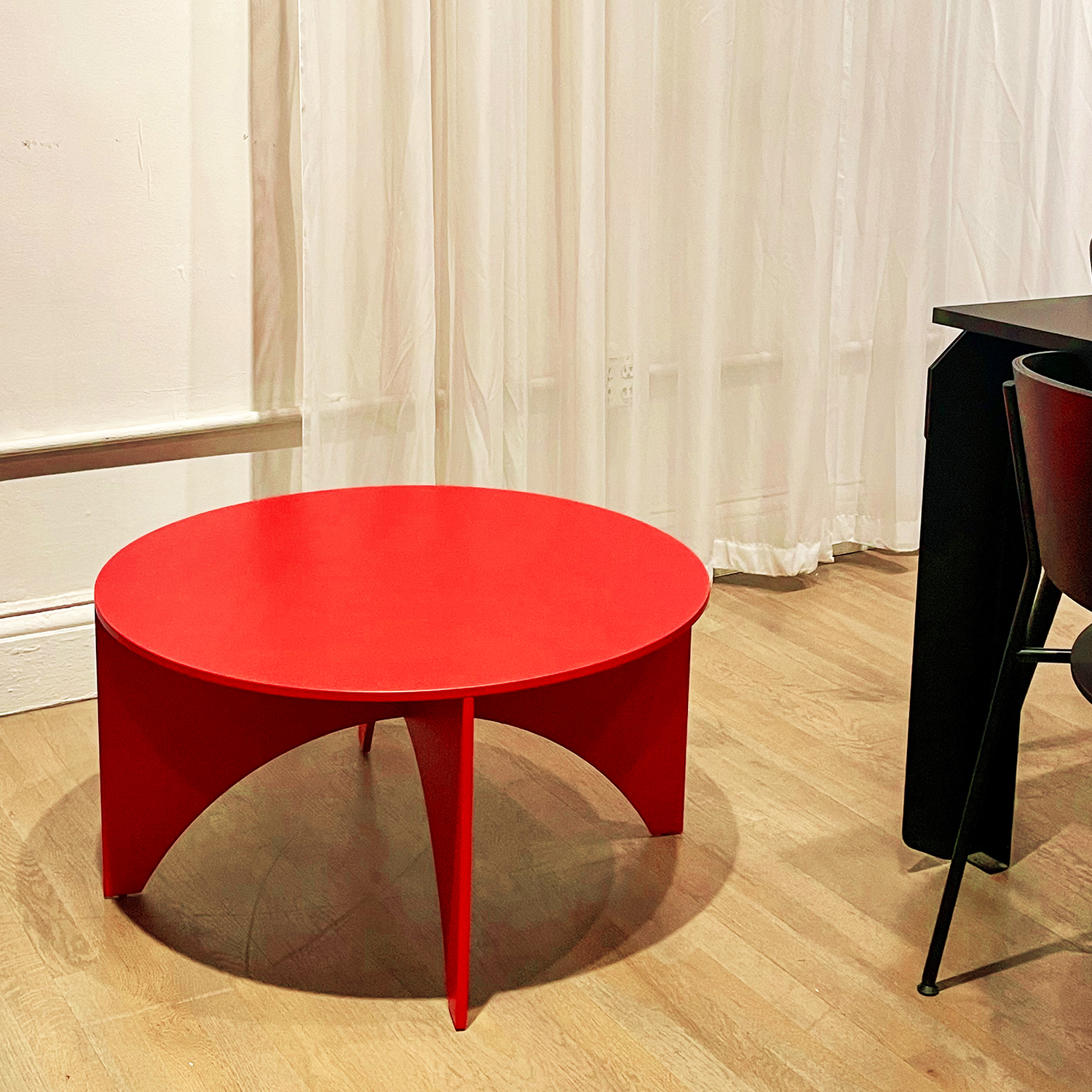 A bright red, round FK05 CHARLOTTE - Sale coffee table by E15 features a minimalist design on a wooden floor beside a black chair and table. Sheer white curtains drape the background wall, creating a modern and stylish setting.