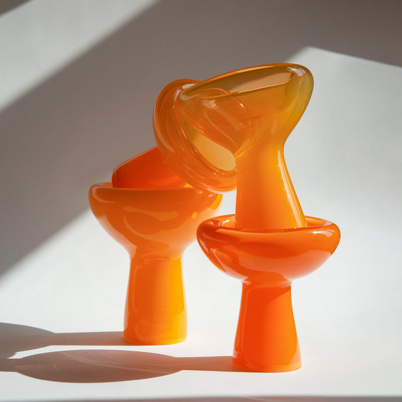 Two glossy, curved orange sculptures with smooth surfaces, echoing the distinctive style of Ursula Futuras PHANTOM Dish, are artistically stacked against a neutral backdrop, creating soft shadows.