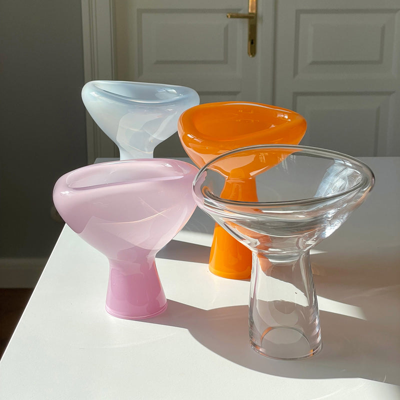 Four PHANTOM Dish vases by Ursula Futura, featuring pastel blue, pink, vibrant orange, and clear glass hues, are arranged on a white surface. Sunlight casts soft shadows as a door with a golden handle highlights their unique design in the background.