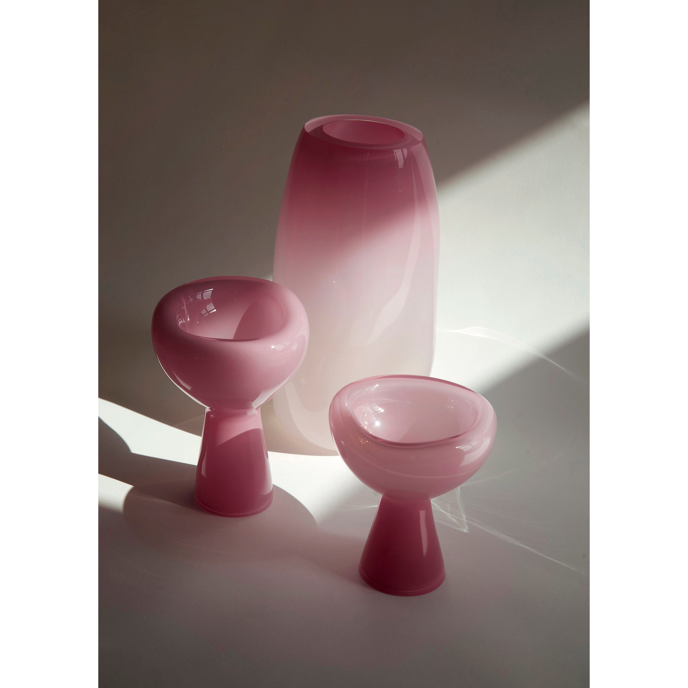 Three PHANTOM Dish pieces by Ursula Futura, crafted from pink mouth-blown glass in varying sizes and shapes, are softly illuminated against a neutral backdrop. The unique design casts gentle shadows on the surface, creating an ethereal ambiance.