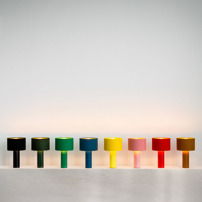 Eight vibrant table lamps, featuring a Victor Foxtrot ALL ROUND MINI with a wool fabric shade, line up against a white wall. The shades and bases in black, dark green, light green, turquoise, yellow, bright yellow, pink, and red create a soft glow.