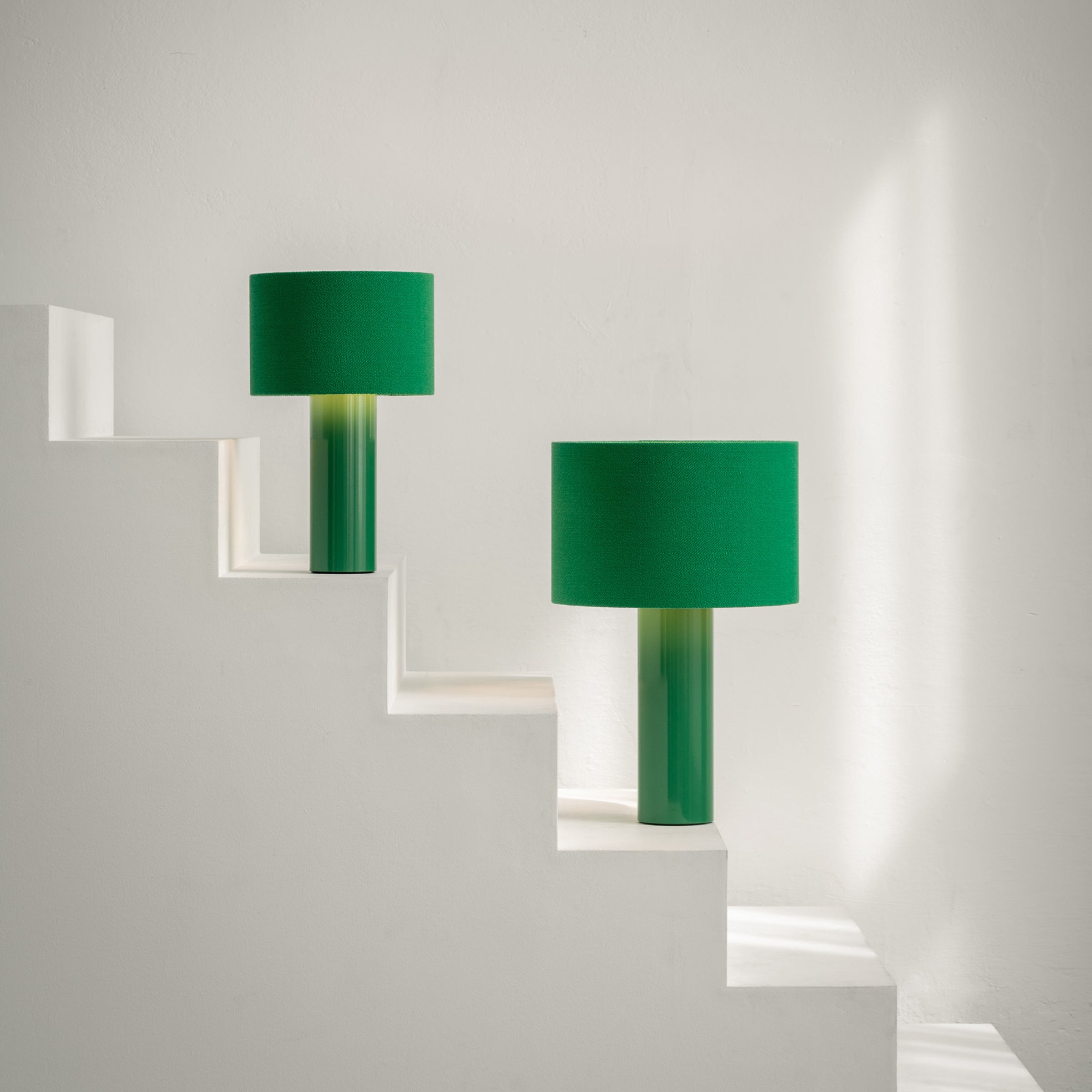 Victor Foxtrots ALL ROUND table lamps, with wool fabric shades and powder-coated steel bases, adorn the ascending white staircase steps. Sunlight casts gentle shadows on the minimalist white wall, crafting a serene and modern ambiance.