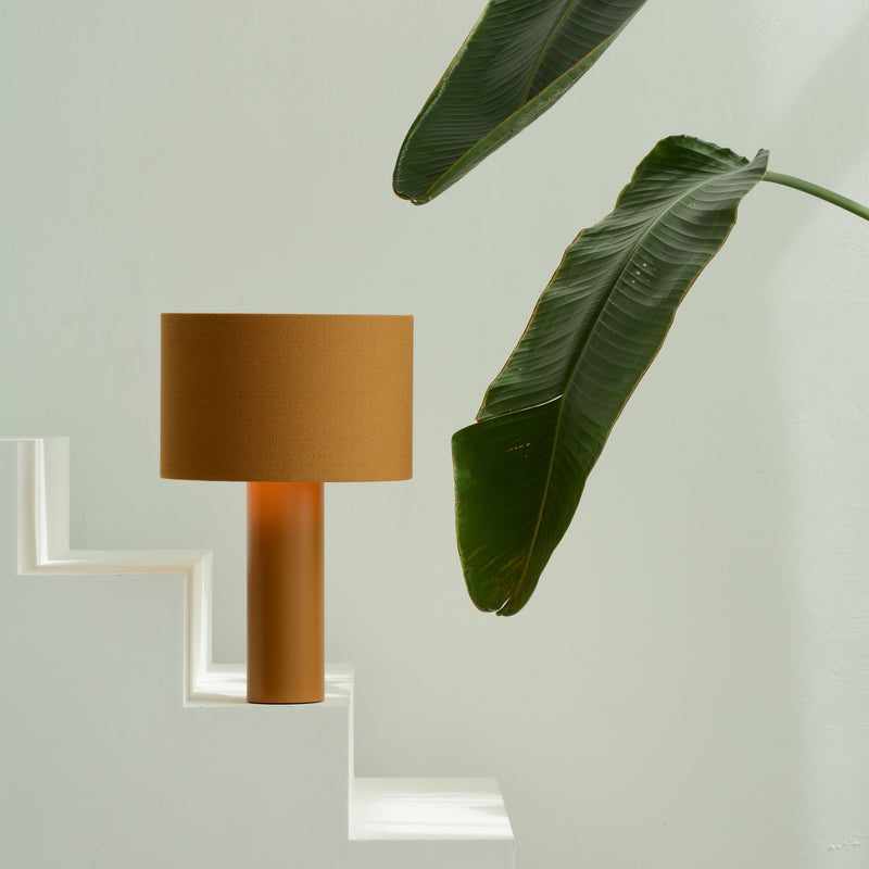 The Victor Foxtrot ALL ROUND Table Lamp, with a modern cylindrical design and wool fabric lampshade, is positioned on a white stepped surface. A large green leafy plant extends diagonally from the top right, set against a light gray background.