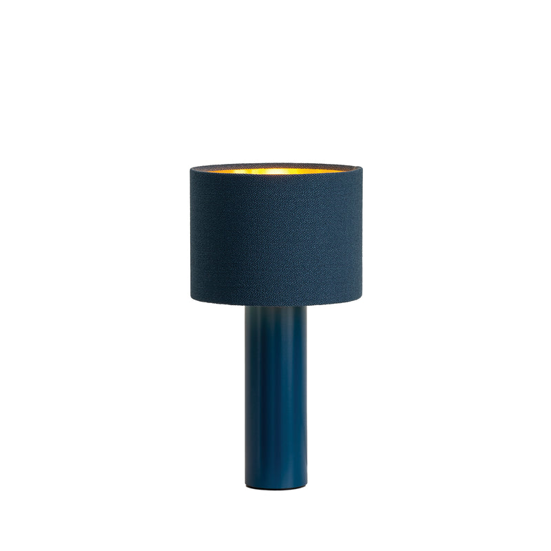 The Victor Foxtrot ALL ROUND MINI Table Lamp is a stylish piece with a blue cylindrical base and matching wool fabric lampshade. It features a gold interior that casts a warm glow, beautifully contrasted against its white backdrop.
