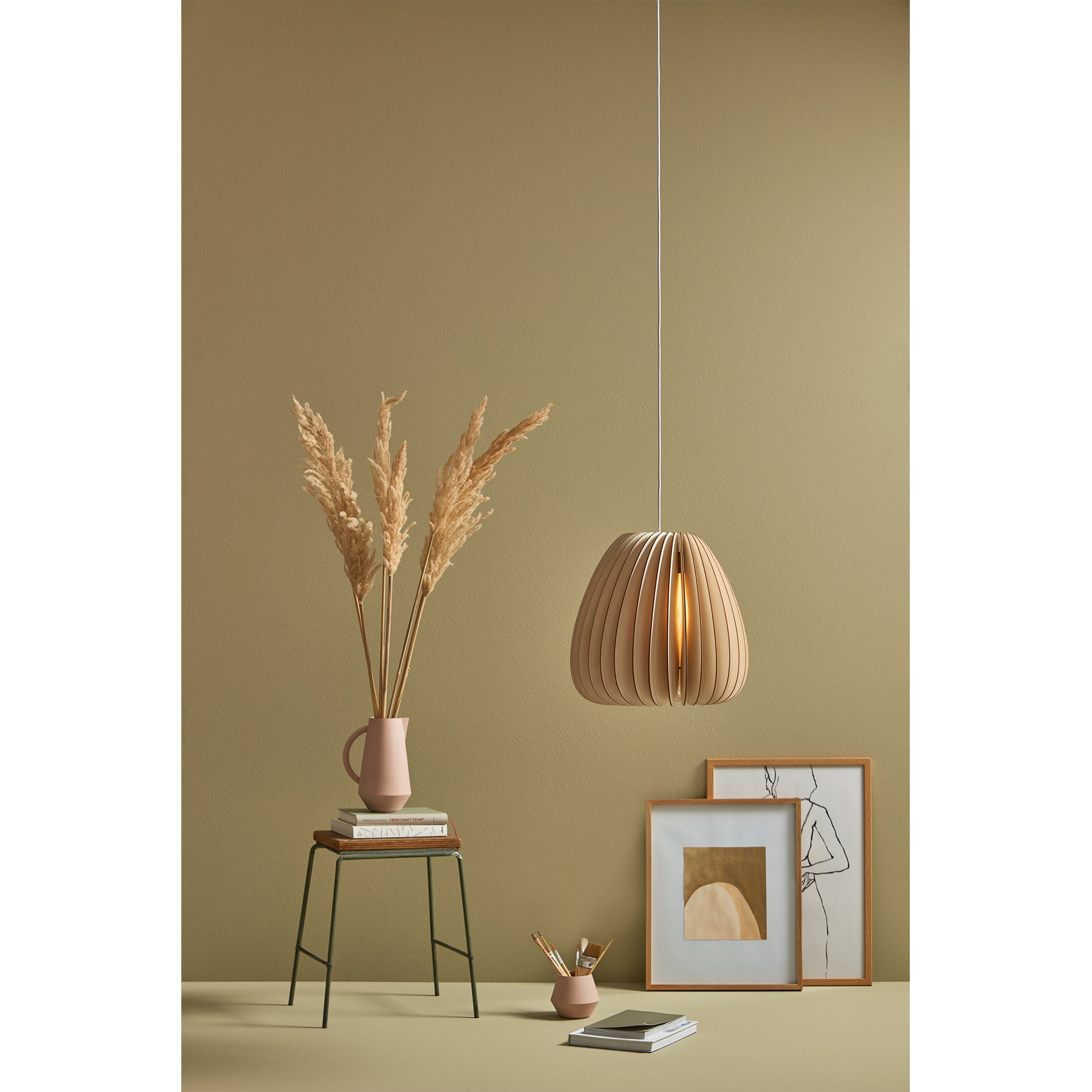 A minimalist room with a tan wall exudes natural ambiance, featuring a Schneid Volum pendant light, a small table with a pink vase of dried pampas grass, and stacked books. Framed abstract art leans against the wall, enhancing the decors organic form. Nearby is a cup filled with pencils.