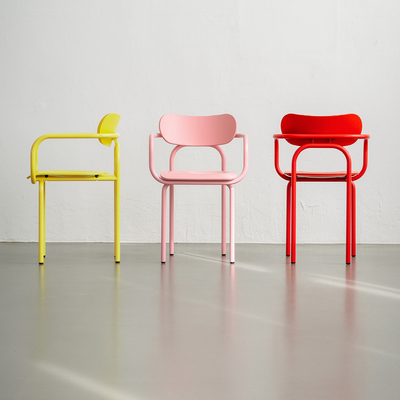 In a minimalist space, three vibrant chairs stand out: a yellow chair facing left, Victor Foxtrots pink Double U Armchair facing forward, and a red chair viewed from behind. Each piece displays iconic aesthetics on a gray floor against a white wall.