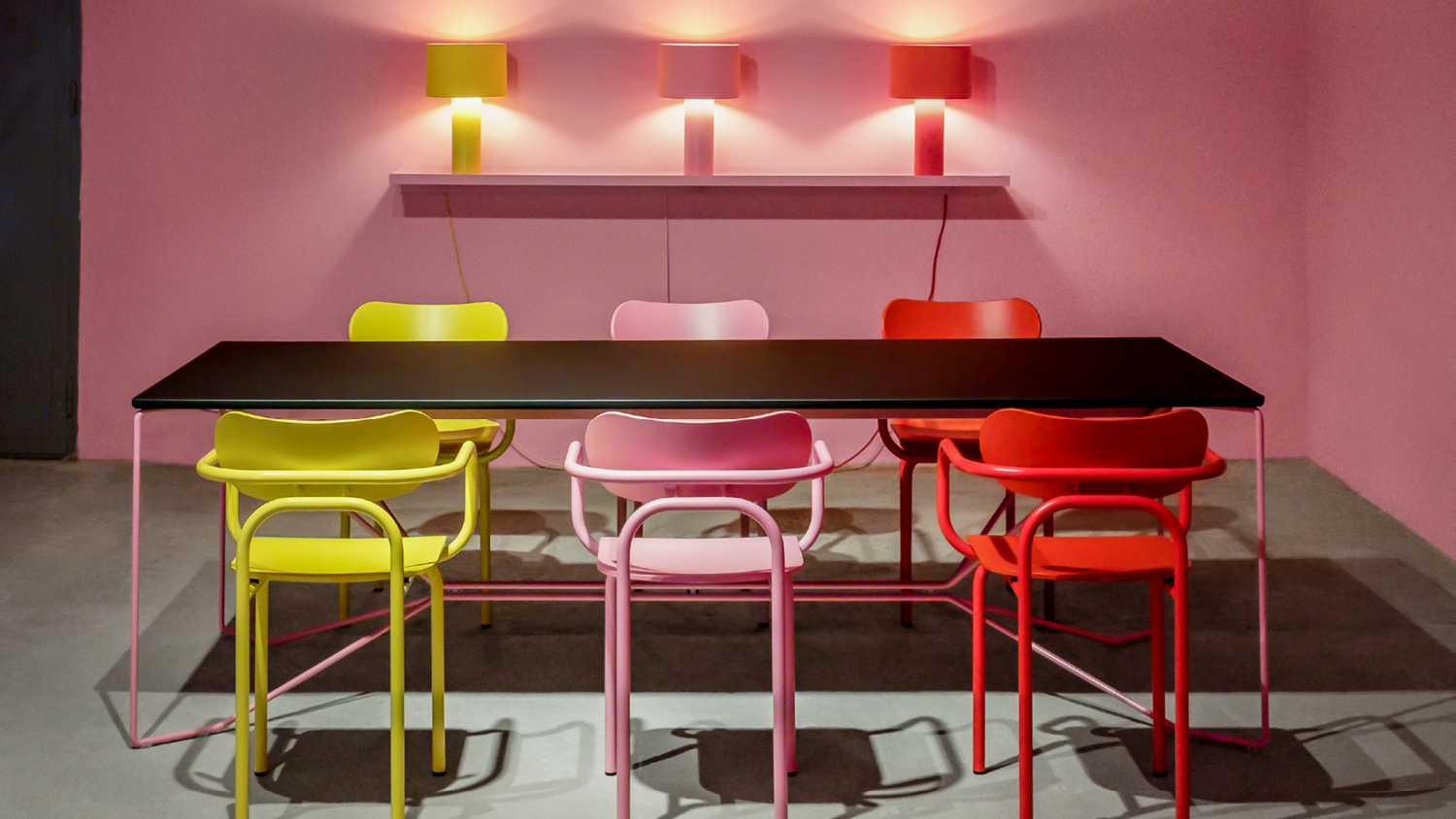 A modern room with a pink wall displays iconic aesthetics, featuring a long black table surrounded by vibrant Victor Foxtrot Double U Armchairs in yellow, pink, red, and orange. Three matching red lamps above the table cast soft light and shadows.