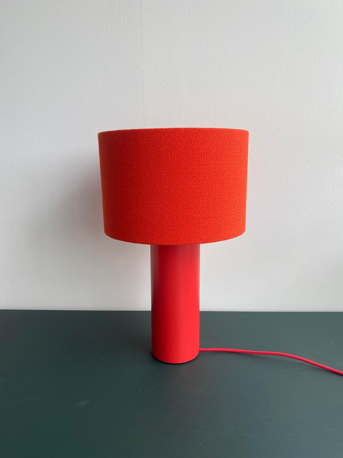 A modern Victor Foxtrot ALL ROUND MINI Table Lamp - SALE, featuring an orange cylindrical base and wool fabric lampshade, is set on a dark green surface against a white background. An elegant orange cord extends from the base, enhancing its sophistication.