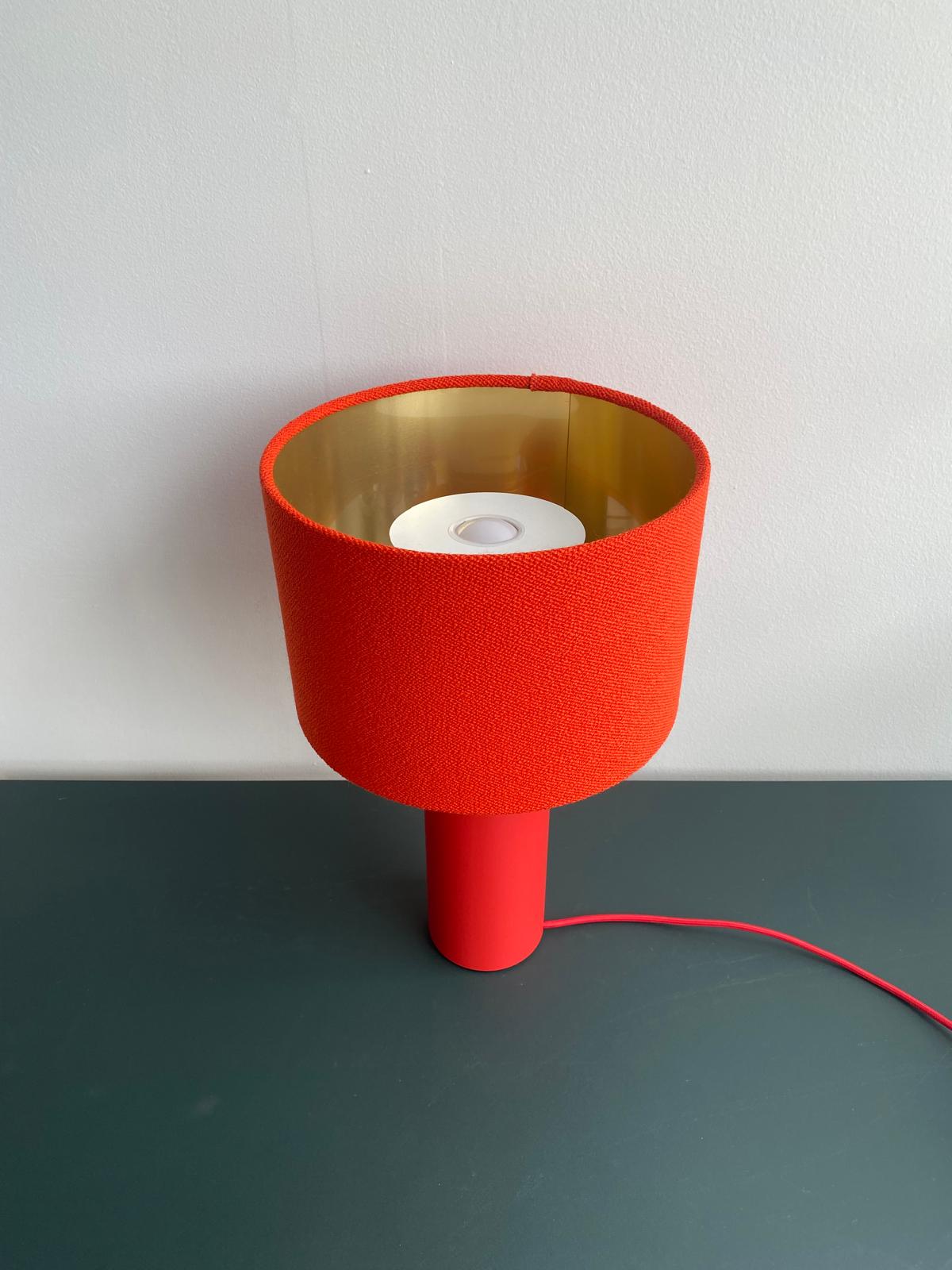 The Victor Foxtrot ALL ROUND MINI Table Lamp - SALE edition, featuring a red cylindrical design with a wool fabric lampshade and gold interior, sits on a dark green surface. A red power cord extends from its base against a plain light-colored background.