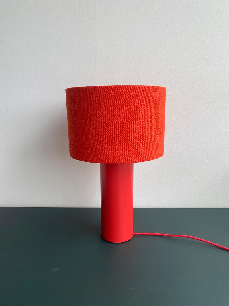 A modern Victor Foxtrot ALL ROUND MINI Table Lamp - SALE, featuring an orange cylindrical base and wool fabric lampshade, is set on a dark green surface against a white background. An elegant orange cord extends from the base, enhancing its sophistication.