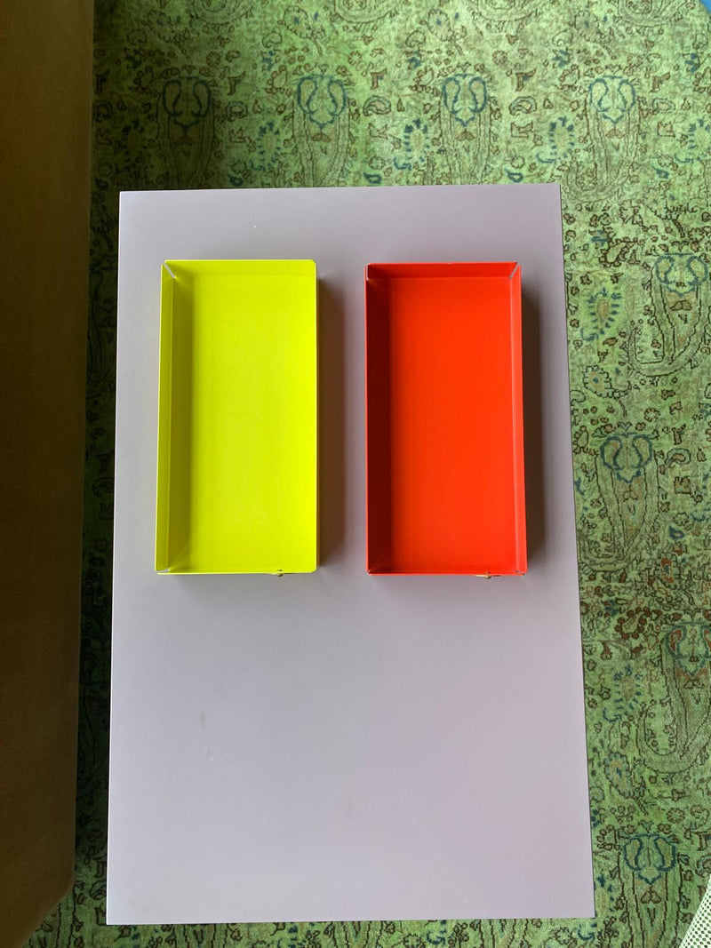 The yellow and red POI Tray M by Victor Foxtrot, made from powder-coated steel, sit side by side on a light gray table adorned with a green patterned carpet, ideal for design enthusiasts.