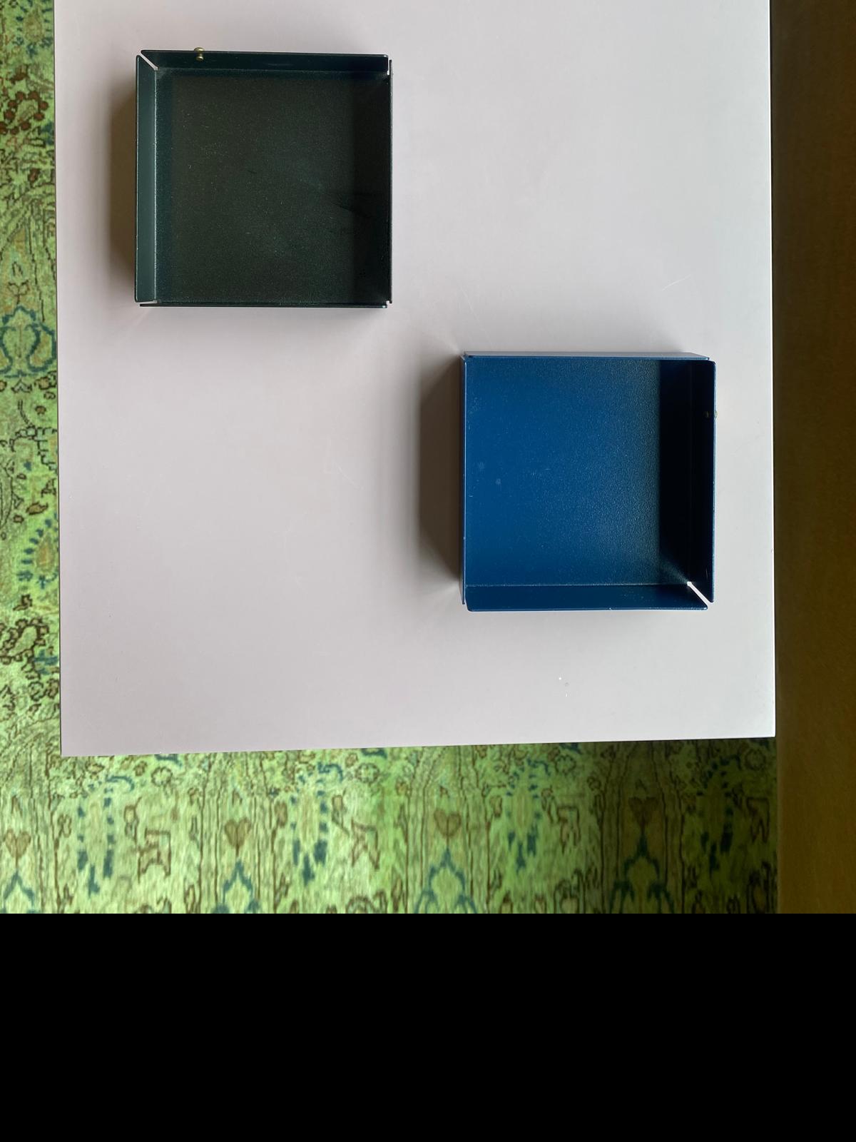 On a white square surface, two Victor Foxtrot POI Tray S - Sale pieces made of powder-coated steel create an elaborate design setting. A black tray is positioned top left and a blue one bottom right against a lush green, patterned background.
