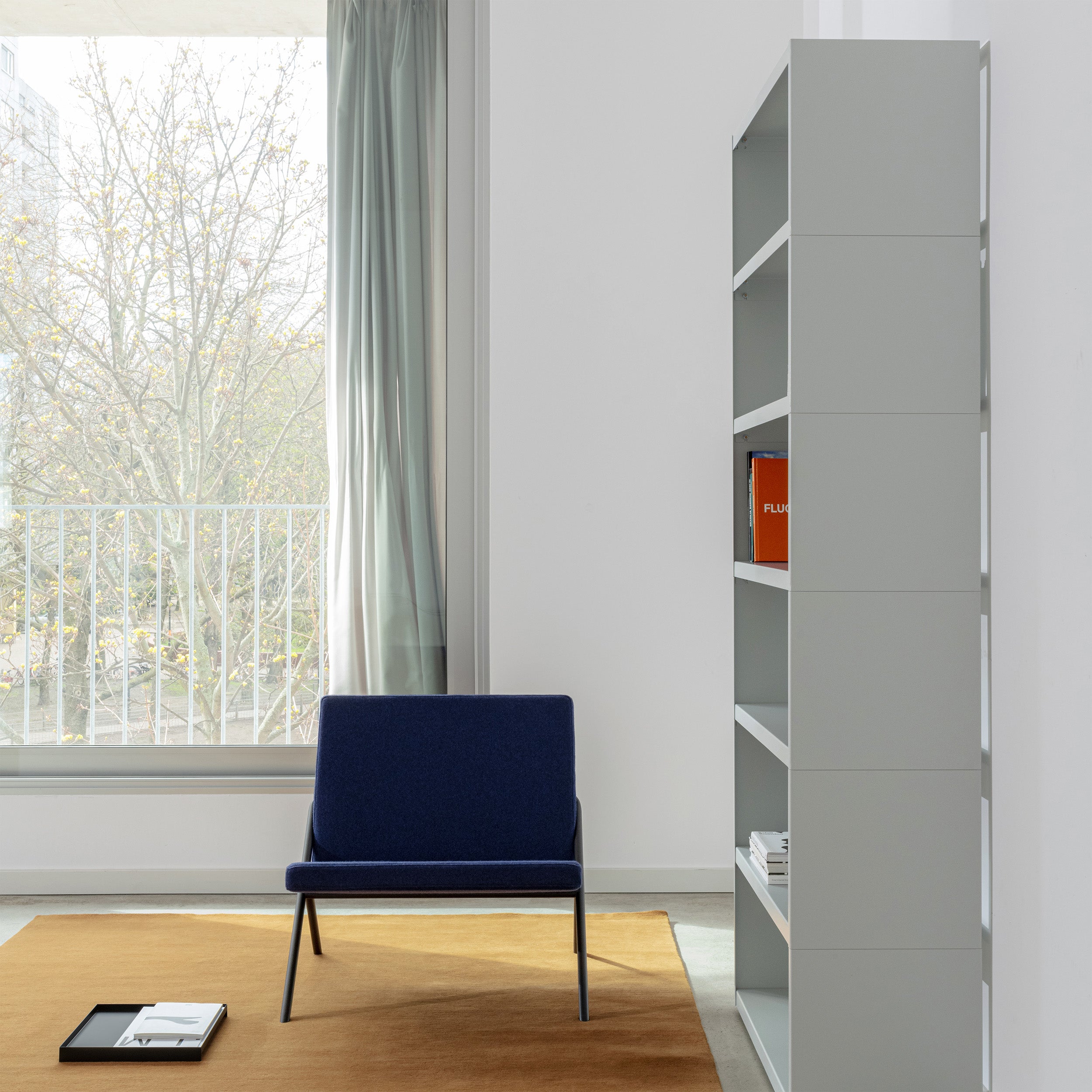 A minimalist room features a blue chair on a brown rug next to the tall W1 Stack Shelves by Loehr, holding just a few items. A large window with sheer curtains invites natural light, revealing the bare trees outside.