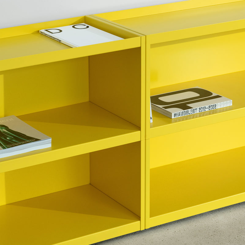 Loehrs W1 Stack Shelves in yellow, featuring a minimalist design, hold several books, including a white one with a black D and HANDBLÆT 2012–2022. These modular units rest on an industrial-style concrete floor against a light gray wall.