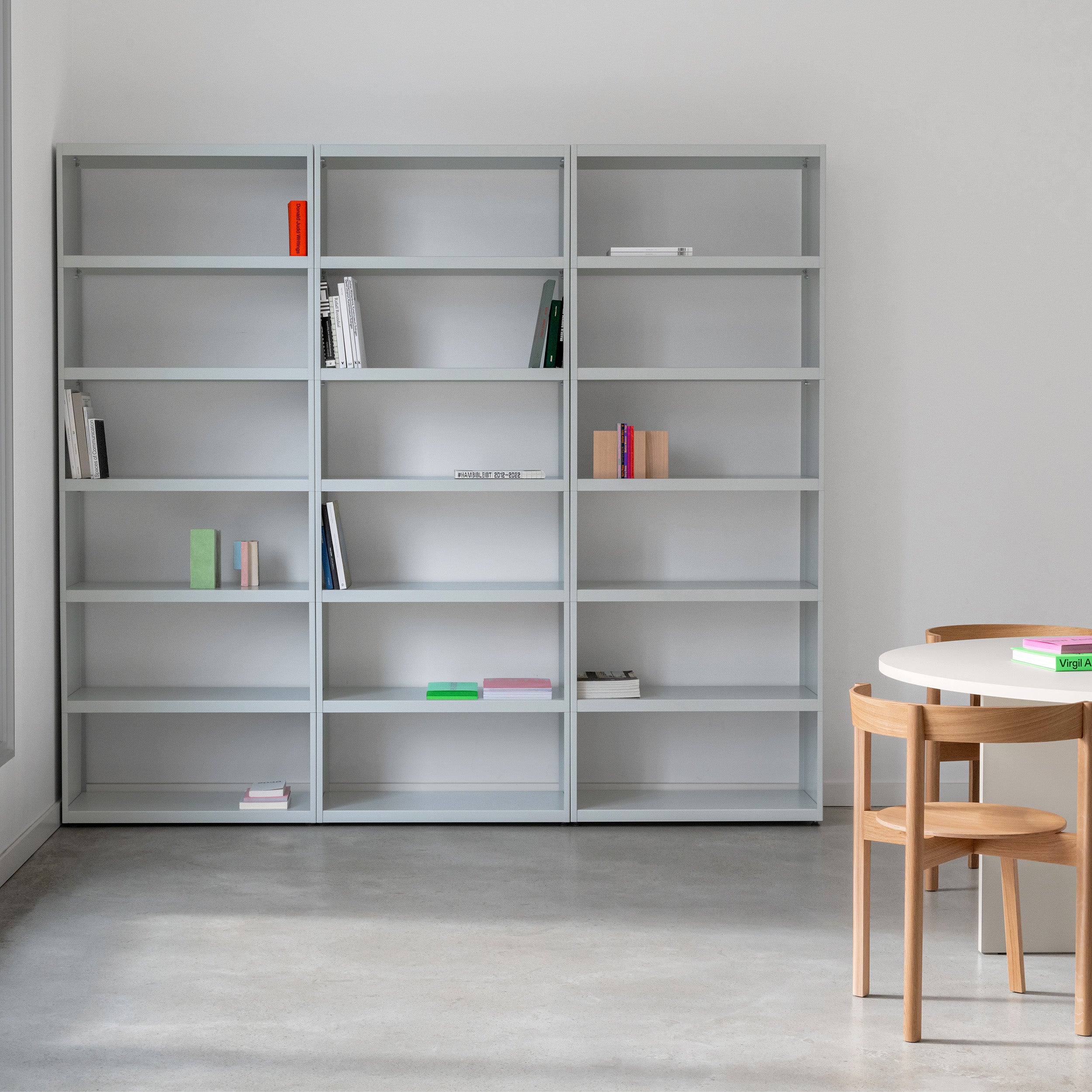 A minimalist room showcases Loehrs W1 Stack Shelves in light gray, partially filled with books. A round white table and two wooden chairs sit on a polished concrete floor. The space is well-lit and uncluttered, highlighting simplicity and modern design with an industrial touch.