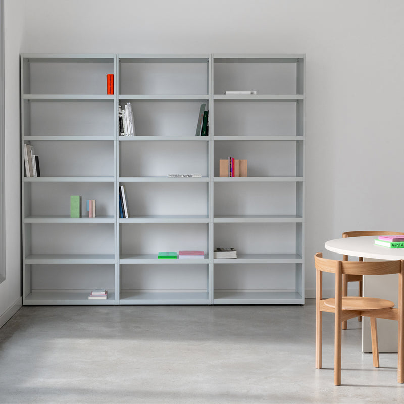 A minimalist room showcases Loehrs W1 Stack Shelves in light gray, partially filled with books. A round white table and two wooden chairs sit on a polished concrete floor. The space is well-lit and uncluttered, highlighting simplicity and modern design with an industrial touch.