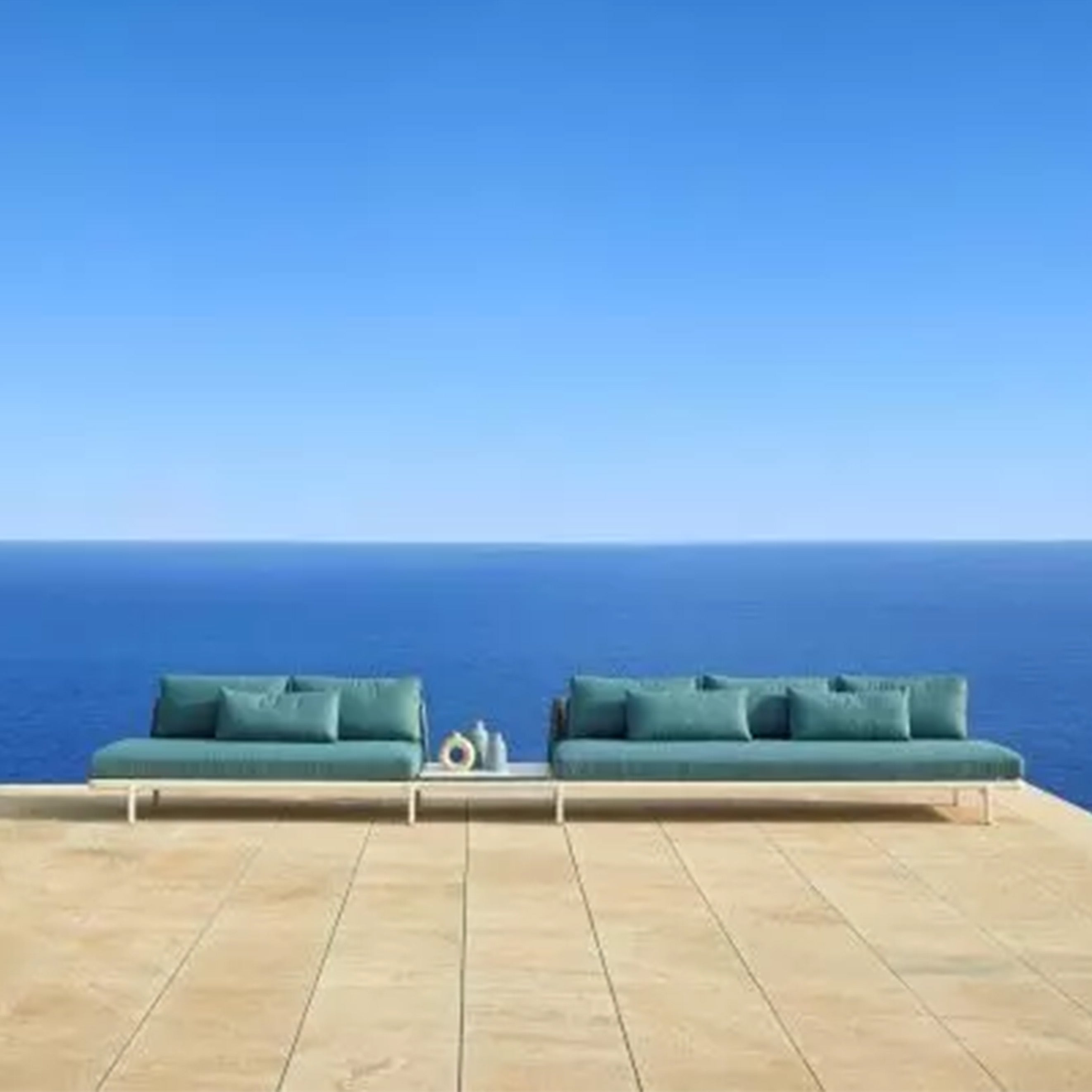 Two teal Resol Anthea Sofa System units with matching pillows adorn a beige tiled terrace by a clear blue ocean under a cloudless sky. Their sleek aluminum frames complement the small table between them, holding decorative items to create a serene and minimalist outdoor setting.