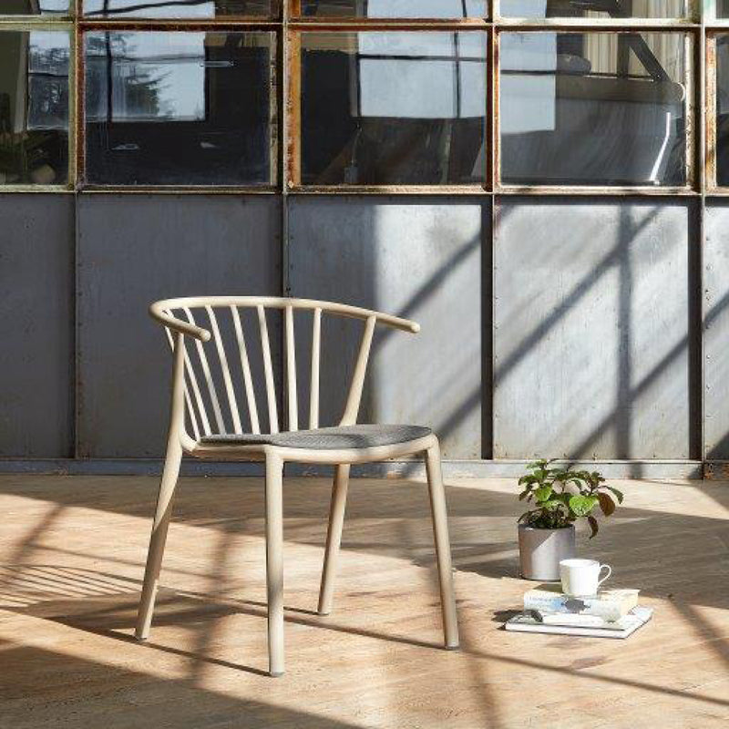 The Resol Woody Upholstered Chair, with its curved backrest, sits on a sunlit wooden floor near a large metal-framed window. This sustainable piece is paired with a small potted plant and a white mug casting shadows in the sunlight.