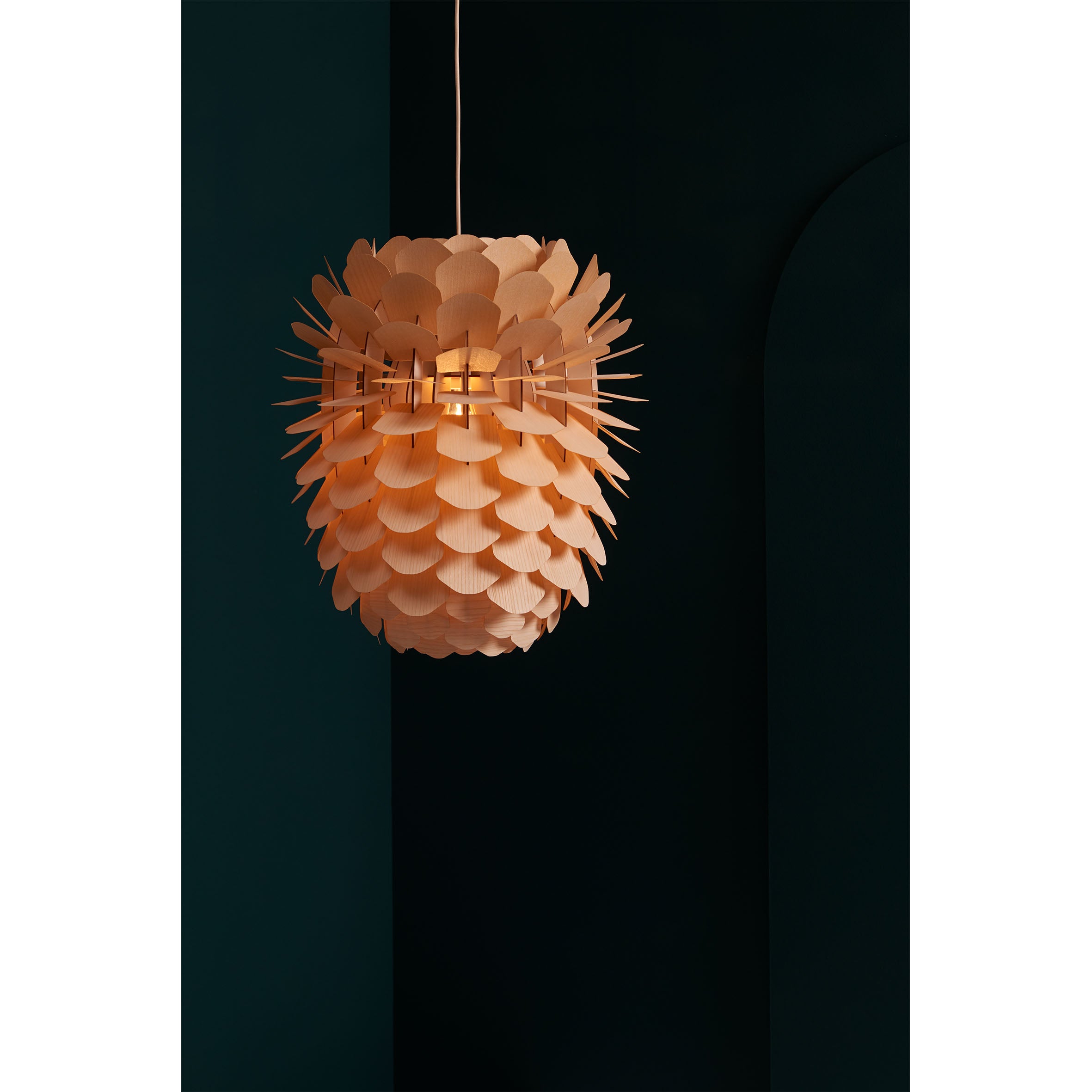 The ZAPPY ASH lamp by Schneid has an ash or oak veneer with a petal-like design, emitting a warm glow. It hangs from the ceiling against a dark green backdrop, showcasing its intricate texture and the soft ambiance of handmade lighting.